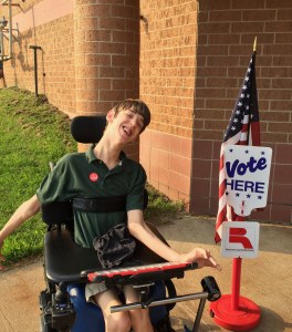 18-year old Justin with I voted sticker on next to Vote Here sign and US Flag