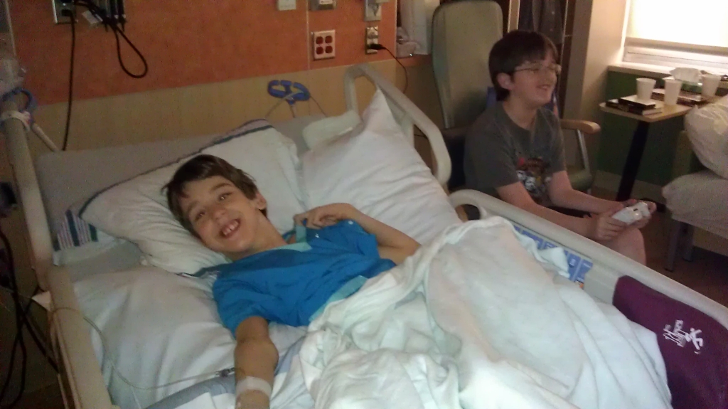 Justin laying in hospital bed, brother sitting in chair with game controller