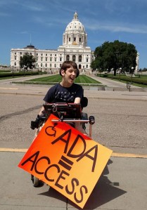Justin in wheelchair holding ADA = Access sign in front of MN State Capitol