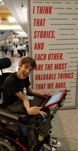 Justin in wheelchair in front of poster that reads "I think that stories, and each other, are the most valuable things that we have." Hank Green