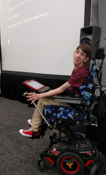 Justin in elevated wheelchair with screen displaying closed captions behind