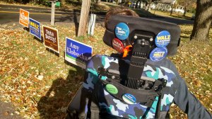 Back of Justin in his wheelchair covered with pins for MN DFL candidates looking at yard signs for DFL candidates