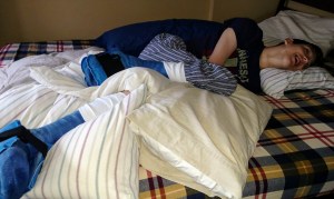 Smiling Justin laying on bed with casts on each leg propped up on pillows