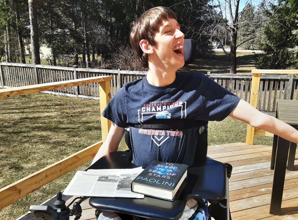 Justin, smiling, seated in power wheelchair on outside deck, To Sleep in a Sea of Stars book by Paolini and newspaper are on lap tray