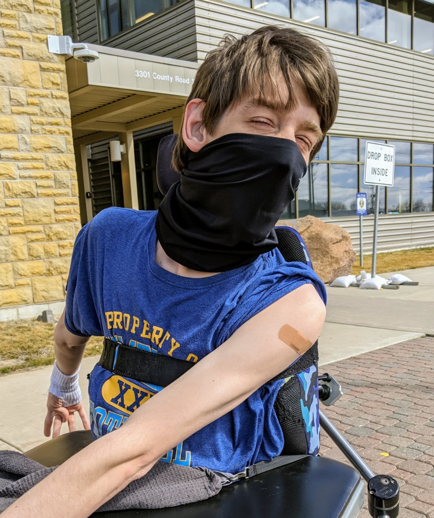 Justin in wheelchair with gator mask on, sleeve rolled up with bandaid on arm, building behind him