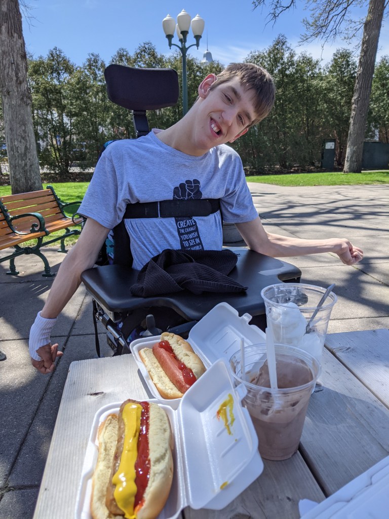 Justin in wheelchair smiling, 2 hot dogs, chocolate and vanilla ice cream in cups on picnic table, trees, park bench
