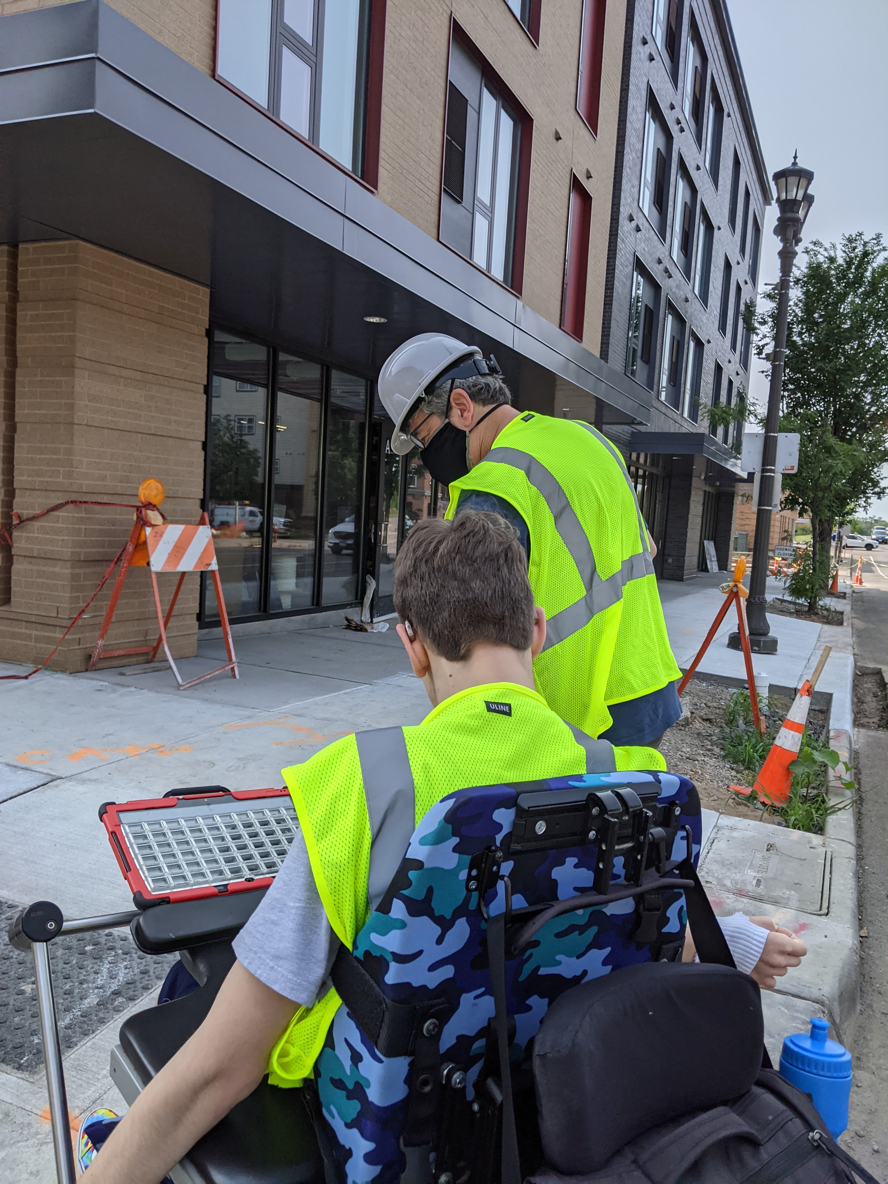Back of masked man with hard hat and yellow vest, back of young man in wheelchair with yellow vest on, apartment building, construction cones