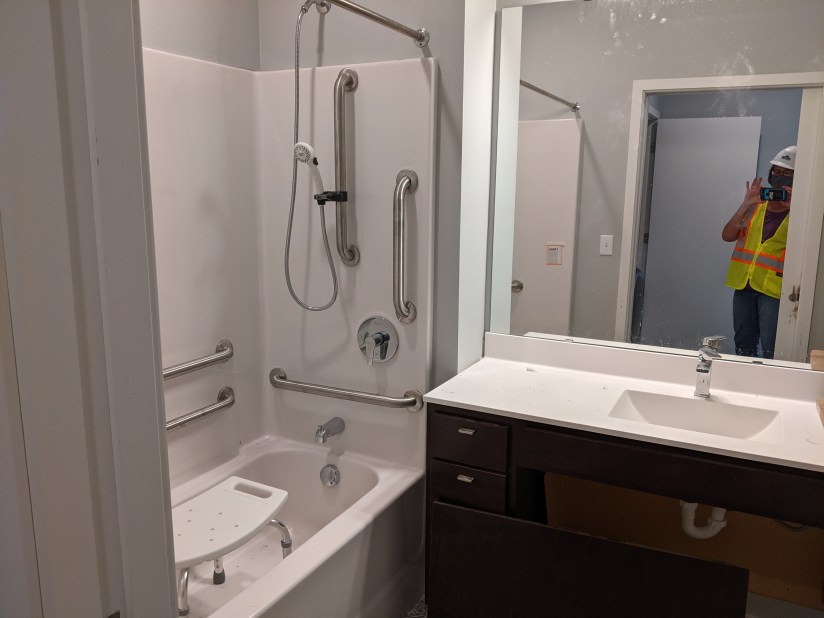 Bathtub with grab bars and shower chair placed lengthwise in tub, woman holding phone with helmet and vest reflected in mirror