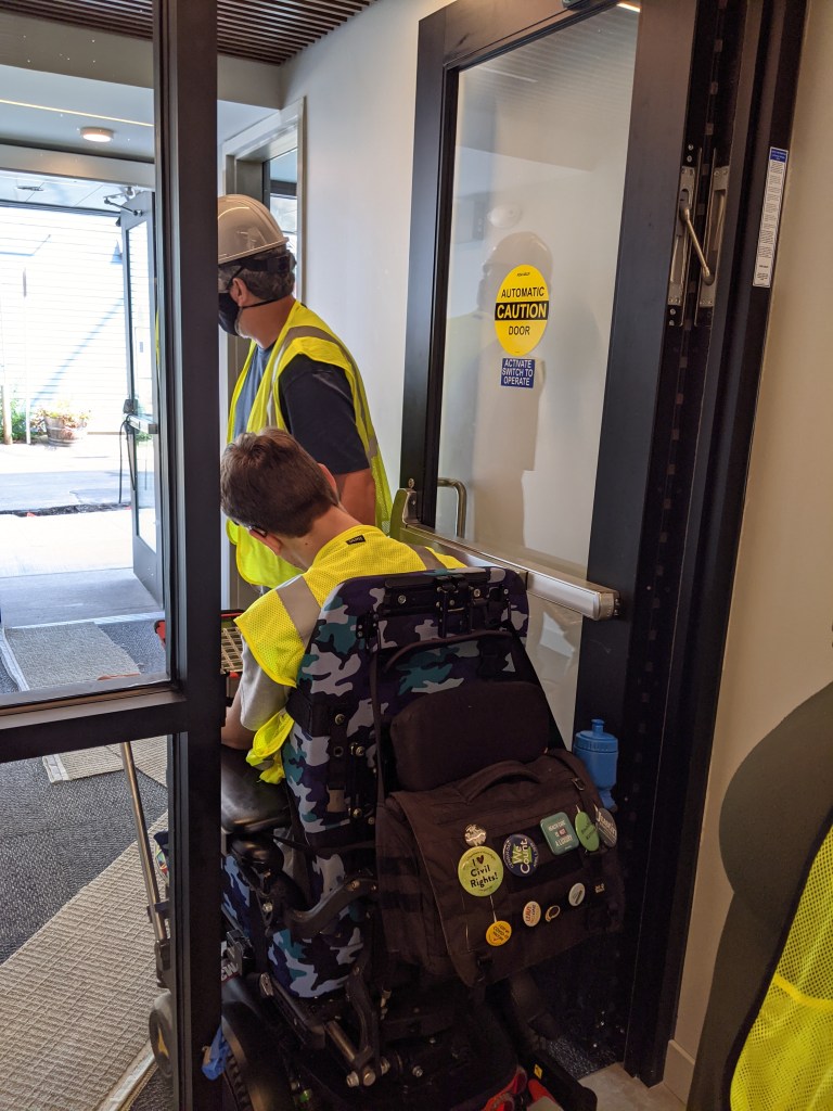 Justin in his wheelchair and his dad exiting doorway, automatic door caution sticker on door