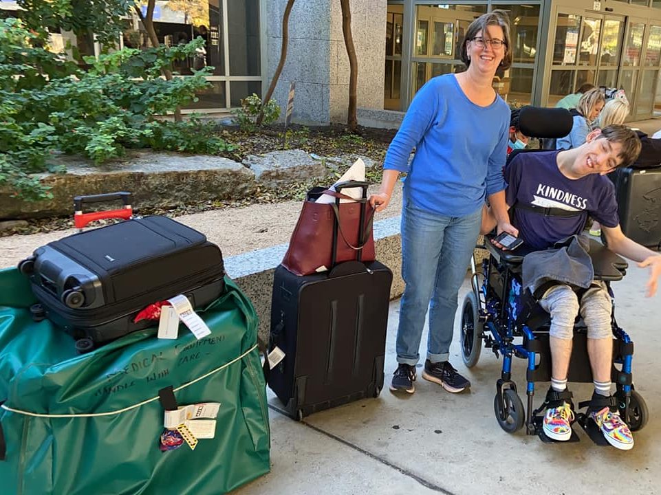 Outside on sidewalk, big green bag, luggage, smiling mom and 23-year old Justin seated in wheelchair
