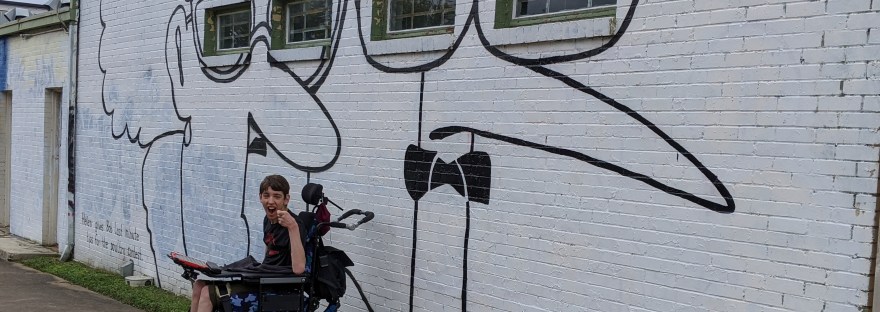 Justin laughing, sitting in wheelchair with communication device, wall mural of cartoon figures of man and woman inspired by Gary Larson