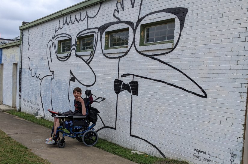 Justin laughing, sitting in wheelchair with communication device, wall mural of cartoon figures of man and woman inspired by Gary Larson