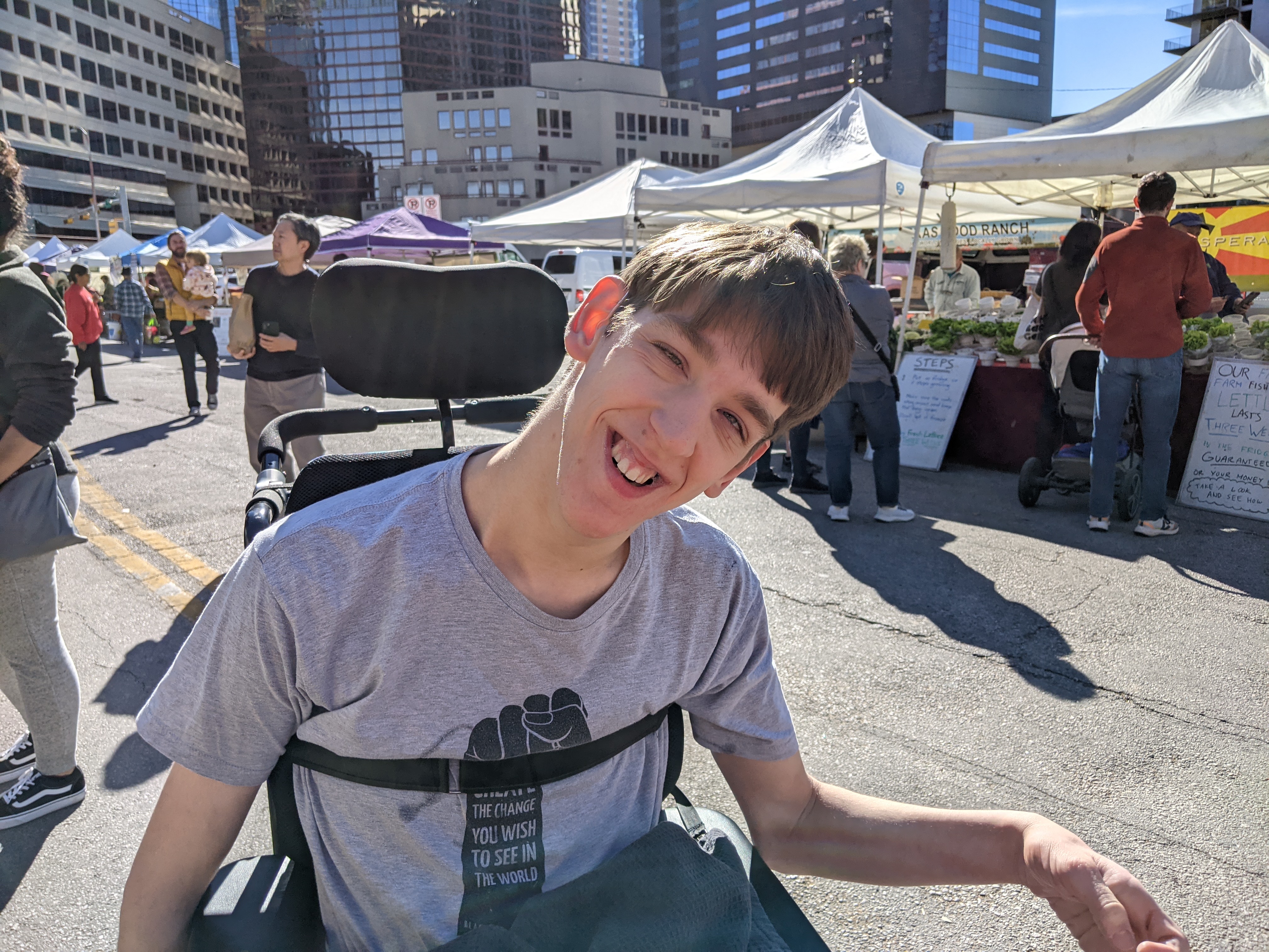 Justin smiling, in wheelchair, Farmer's Market booths on Austin Texas downtown street, people in background, 