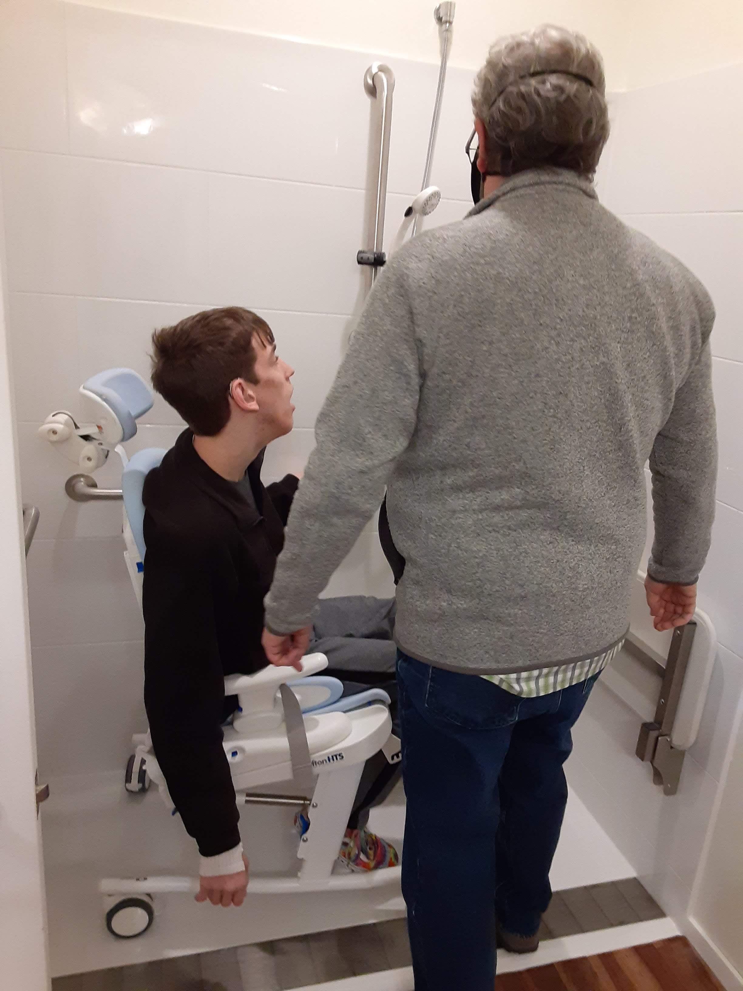 Justin in custom shower chair in roll-in shower, back of his dad standing outside of shower