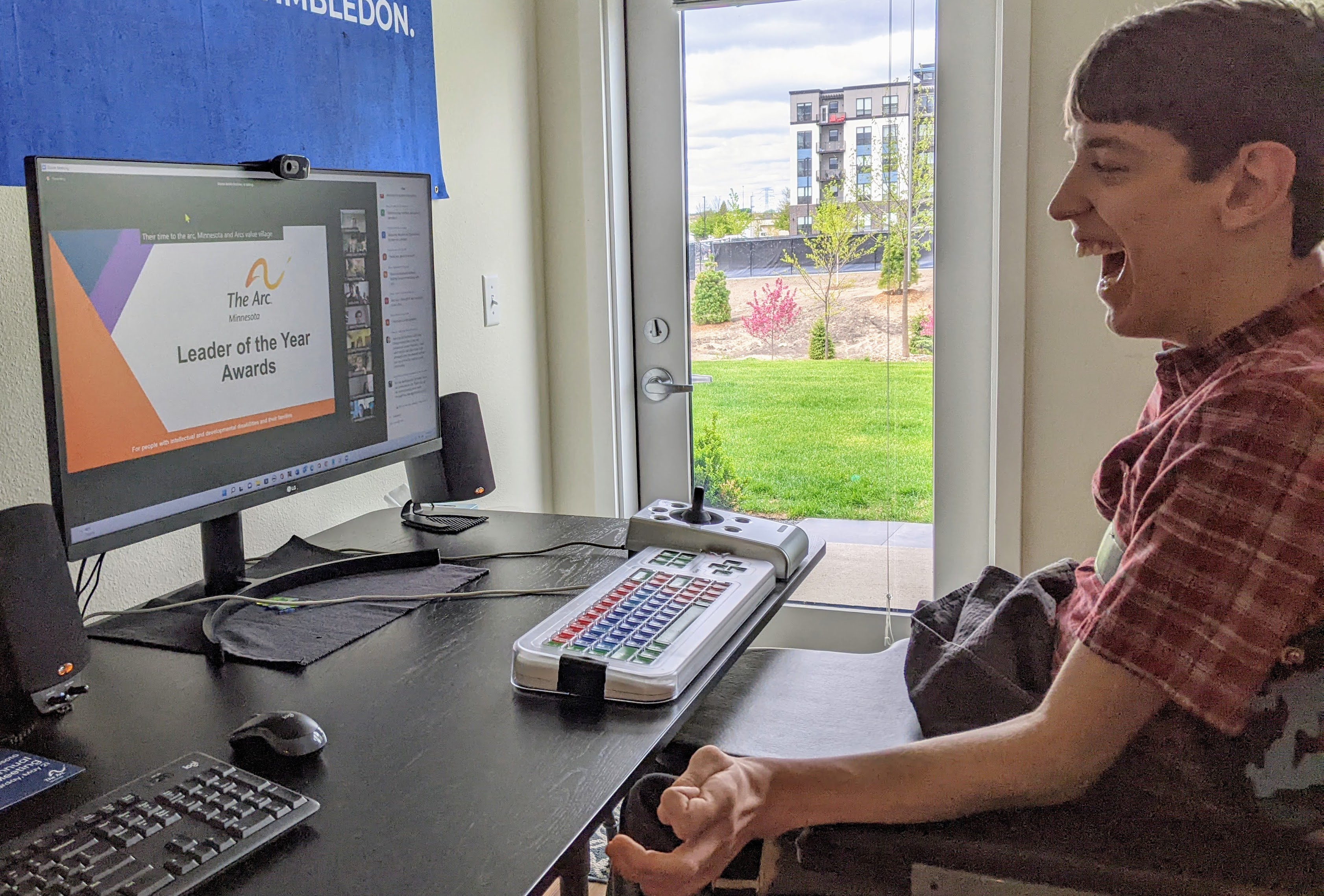 Justin iseated in wheelchair, smiling and looking at computer monitor that says "The Arc Leader of the Year Awards" with web cams and chat for Zoom meeting