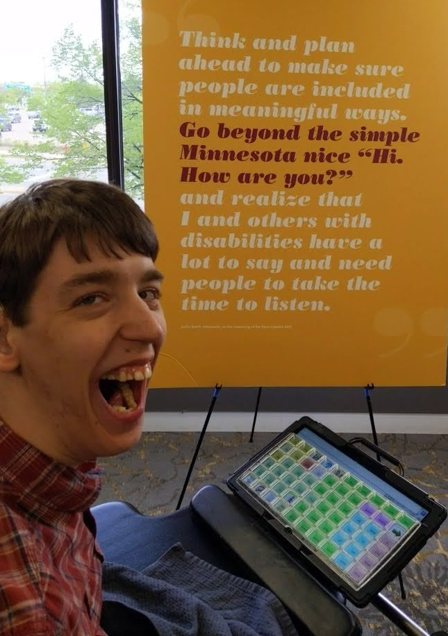 Justin, a smiling young man in wheelchair with communication device, in front of a poster that reads: "Think and plan ahead to make sure people are included in meaningful ways. Go beyond the simple Minnesota nice "Hi, How are you?" and realize that I and others with disabilities have a lot to say and need people to take the time to listen. Quote by Justin Smith, 2017