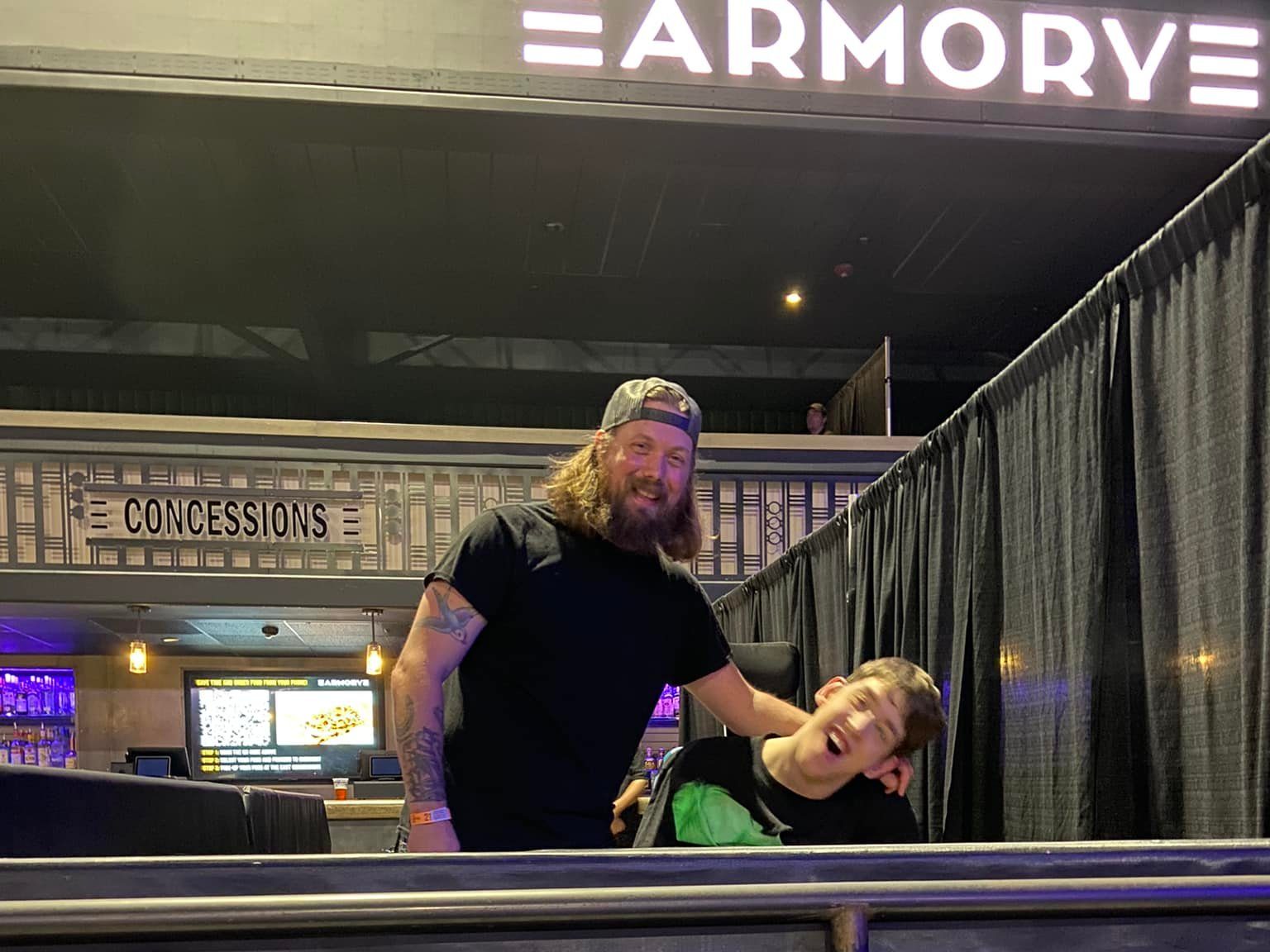 Looking up at Justin and another young man with his arm around him, smiling, Armory sign and concessions behind, curtained off area on the side