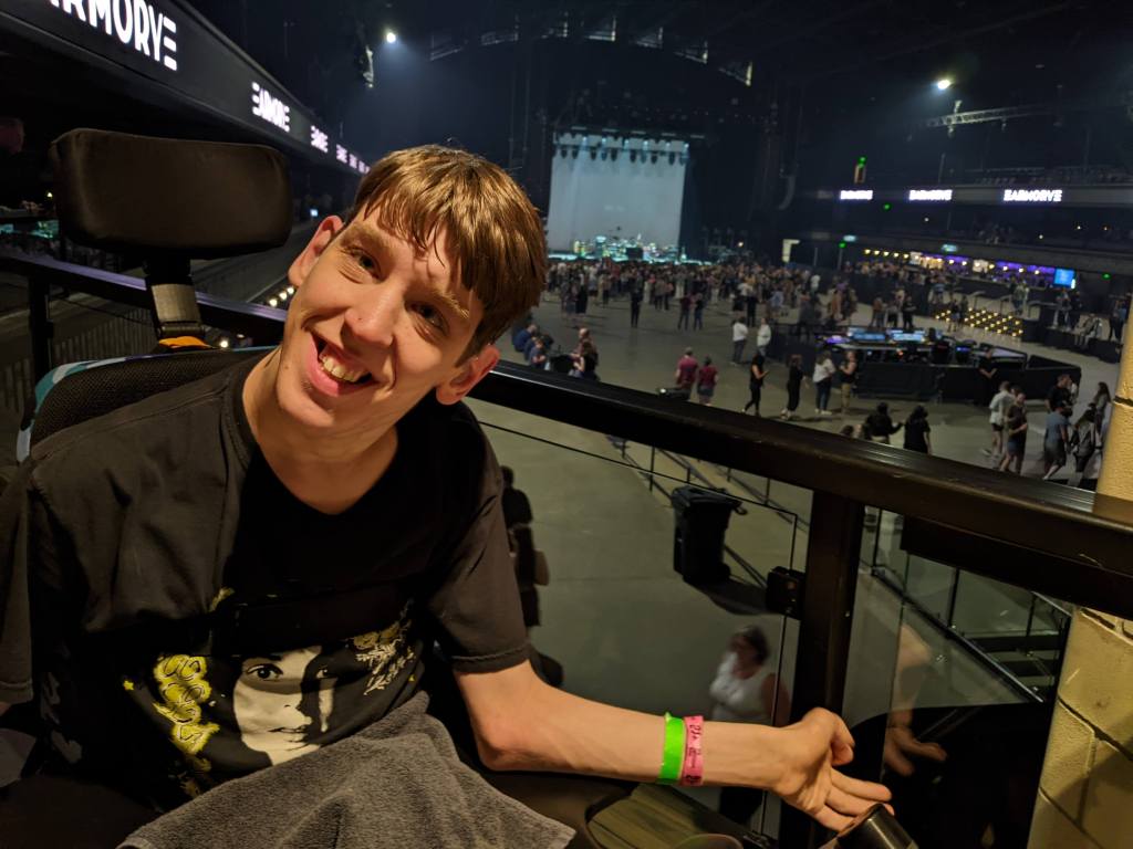 Justin smiling in his wheelchair, 2nd level of Armory in the back looking across people gathering in front of a distant stage