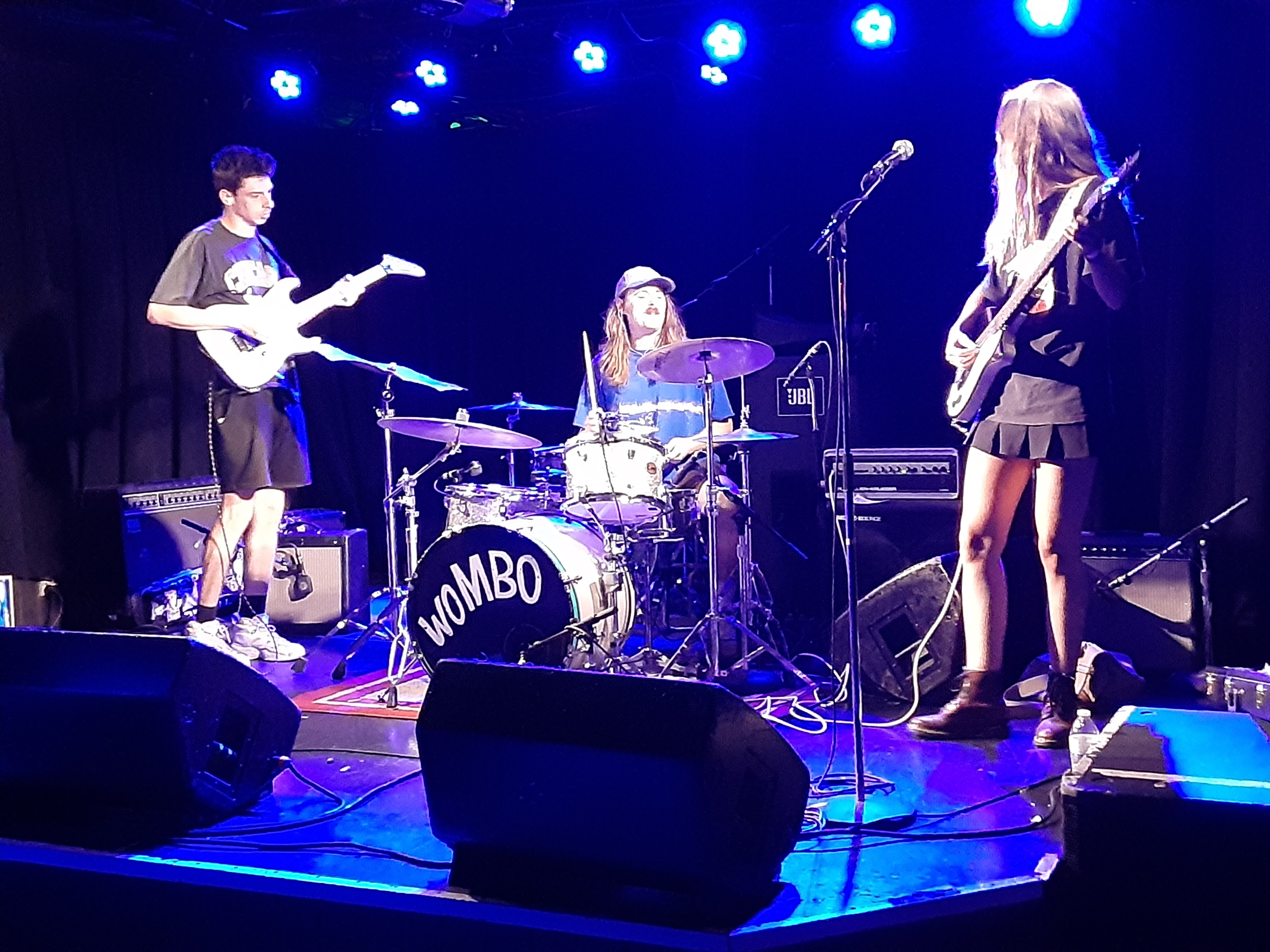 Wombo band with young male guitarist, long hair with mustache drummer, and young woman guitarist on stage