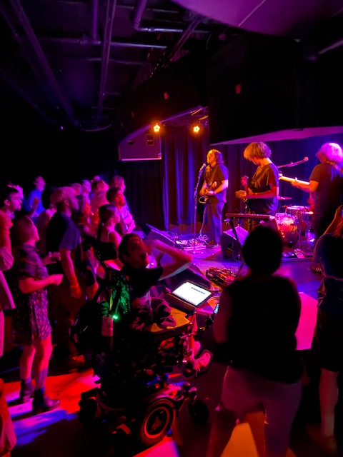 Deeper band with crowd in front of stage, Justin in wheelchair to right of stage