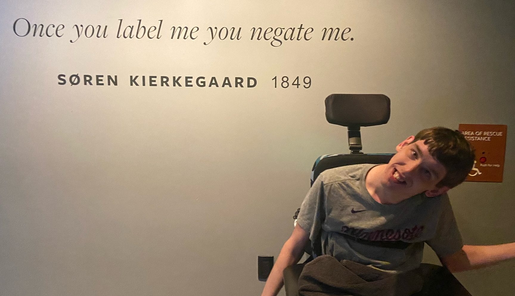 Young man in wheelchair in front of quote painted on wall by Soren Kierkegaard, 1849, "Once you label me you negate me."
