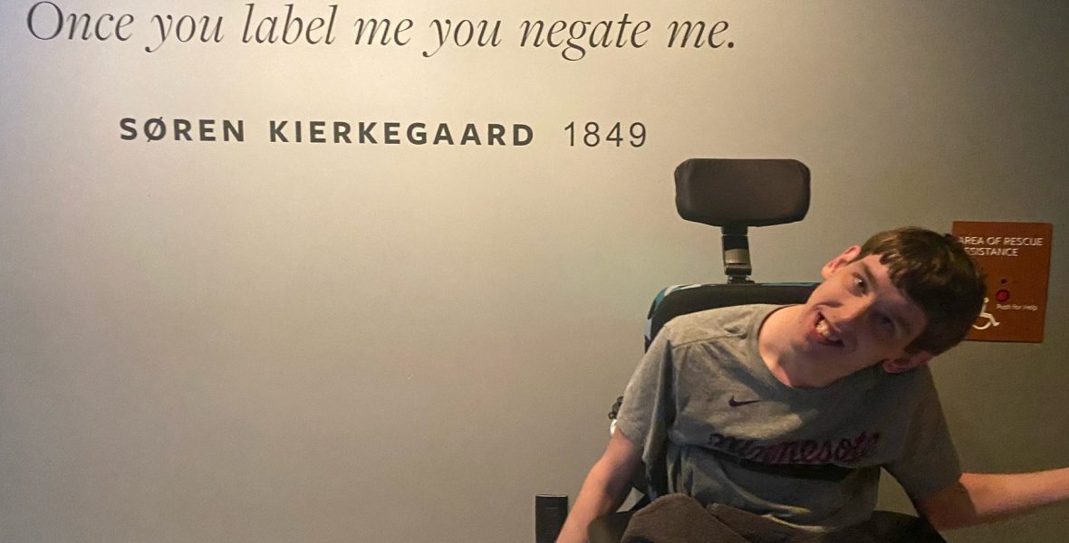 Young man in wheelchair in front of quote painted on wall by Soren Kierkegaard, 1849, "Once you label me you negate me."