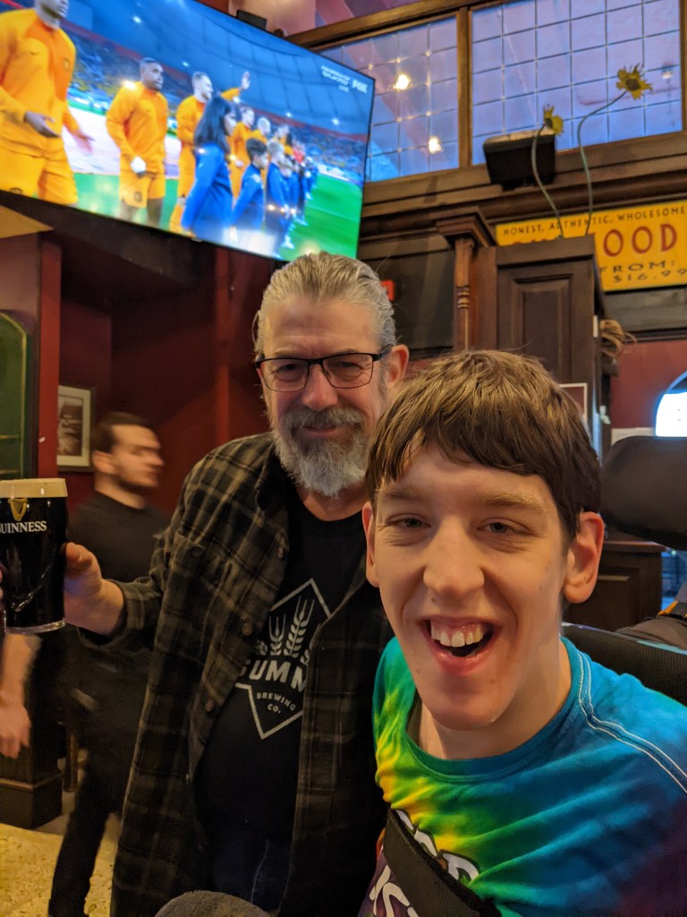 Justin, young man seated in wheelchair, next to his middle-aged dad with grey beard and hair pulled back in ponytail and holding Guinness beer, large screen with football/soccer match, in pub