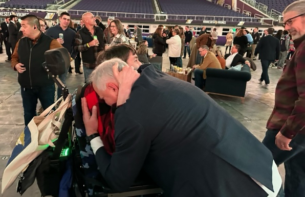 Governor Walz leaning over to hug Justin who is in a power wheelchair, crowd behind