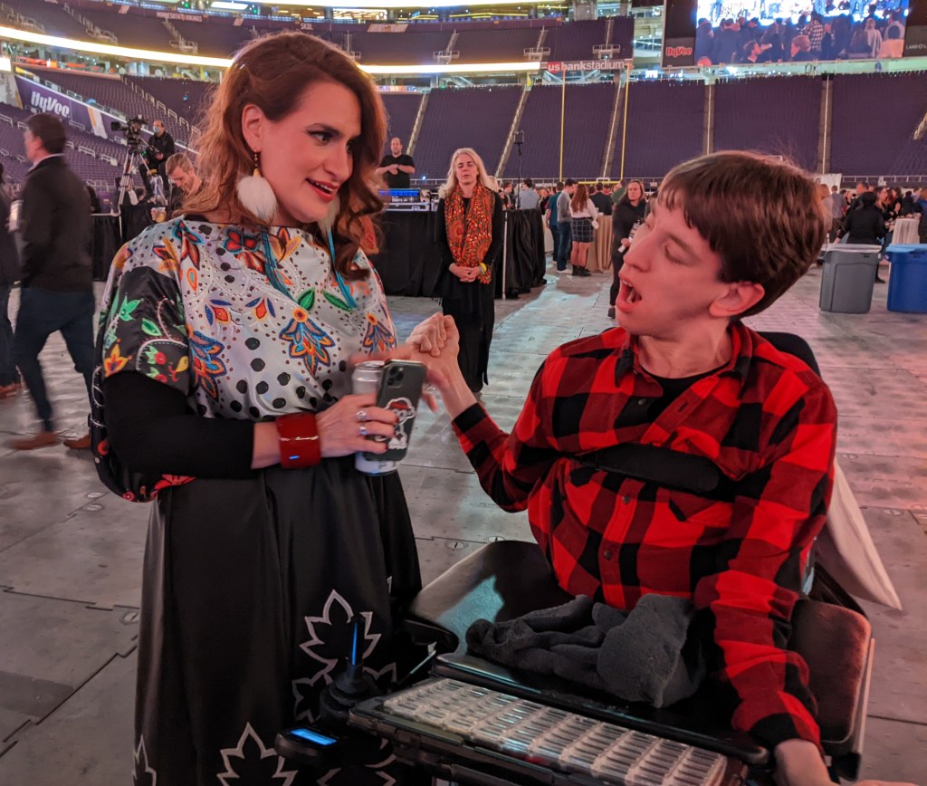 Justin talking to Lieutenant Governor Peggy Flanagan, Justin is a young man in a power wheelchair with communication device