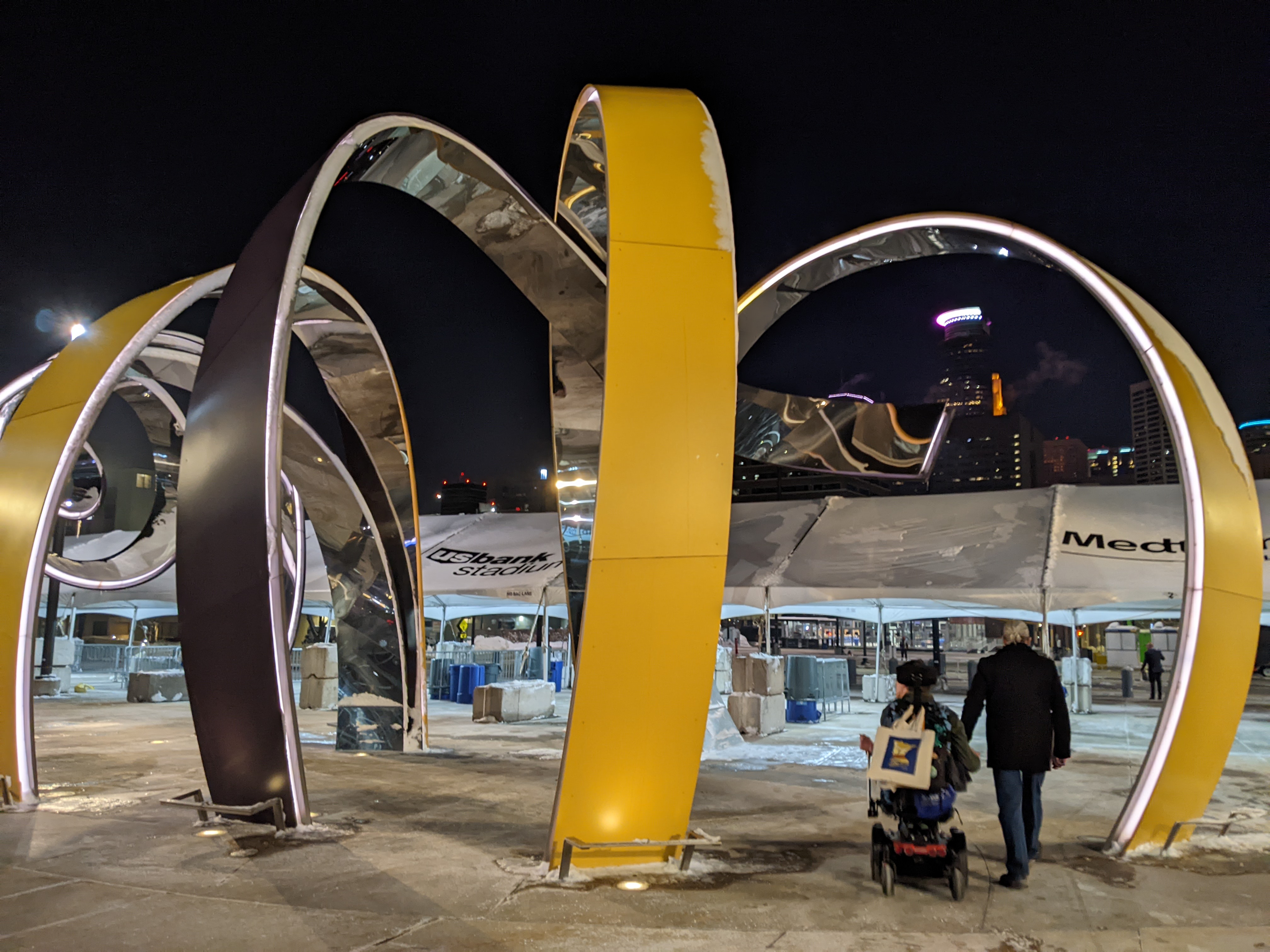 Justin in power wheelchair with One Minnesota bag hanging from back of wheelchair next to his dad, walking under sculpture at US Bank stadium with Minneapolis skyline lit up behind