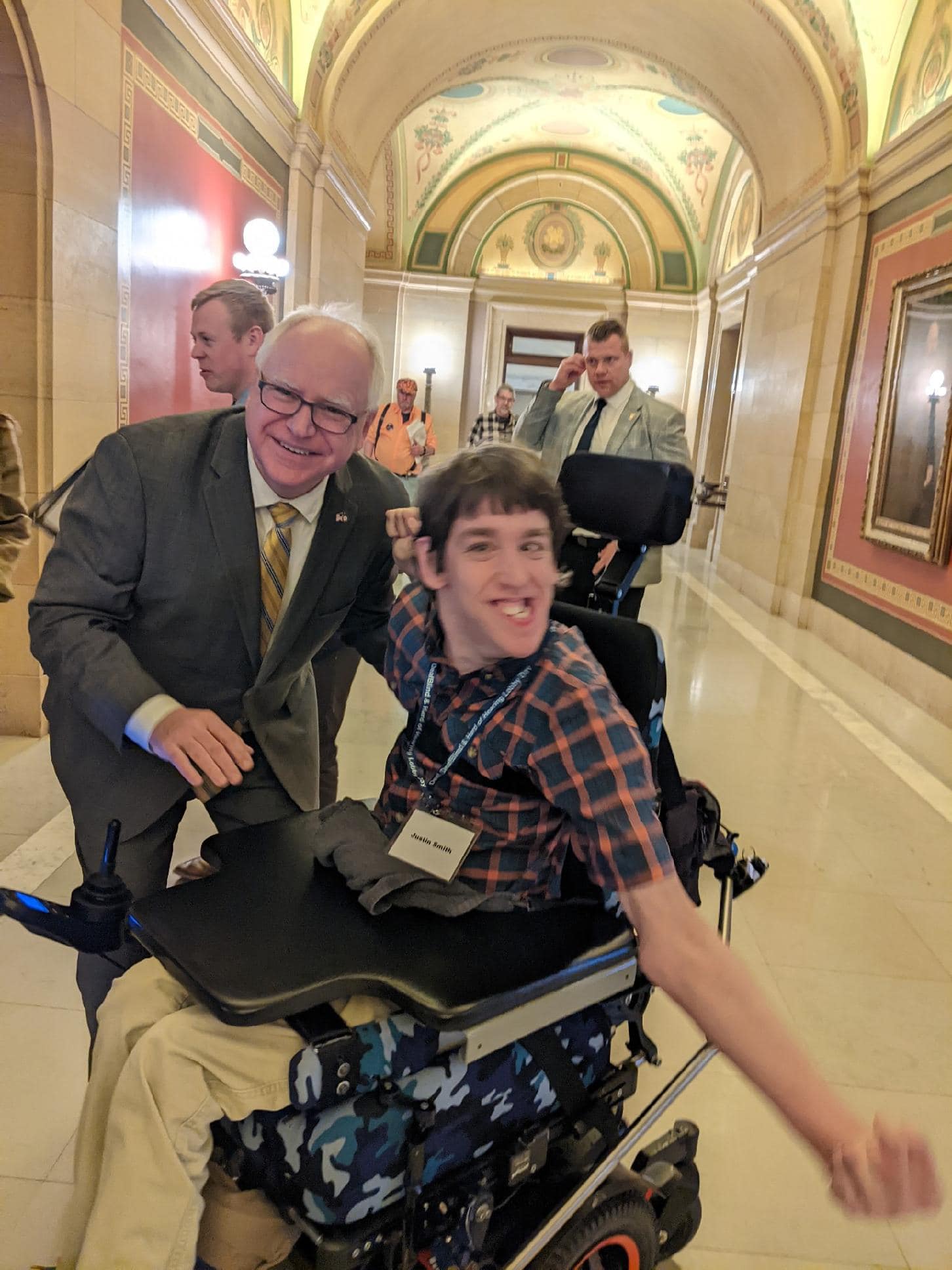 Justin seated in power wheelchair smiling, Governor Walz is leaning next to Justin and smiling, in Capitol hallway with a couple people walking behind