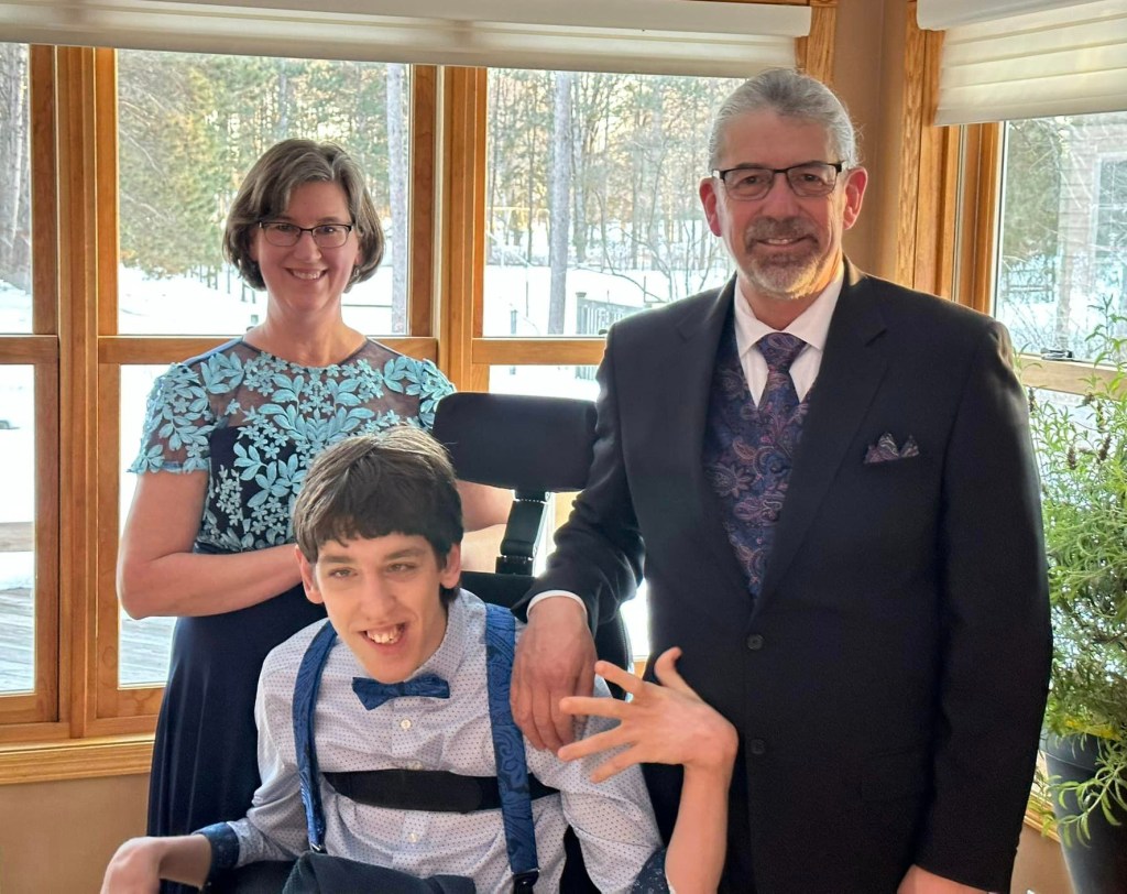 Justin in wheelchair with bow tie, his middle-aged grey-haired dad standing next to him wearing suit, mom is standing behind with dress