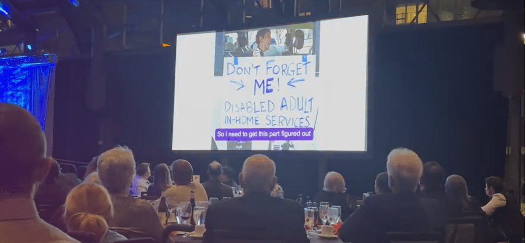 People seated at dining tables watching large screen with picture of Justin in his wheelchair in a minivan. Sign on van says "don't forget me! Disabled adult in-home services." and caption on screen says "So I need to get this part figured out"
