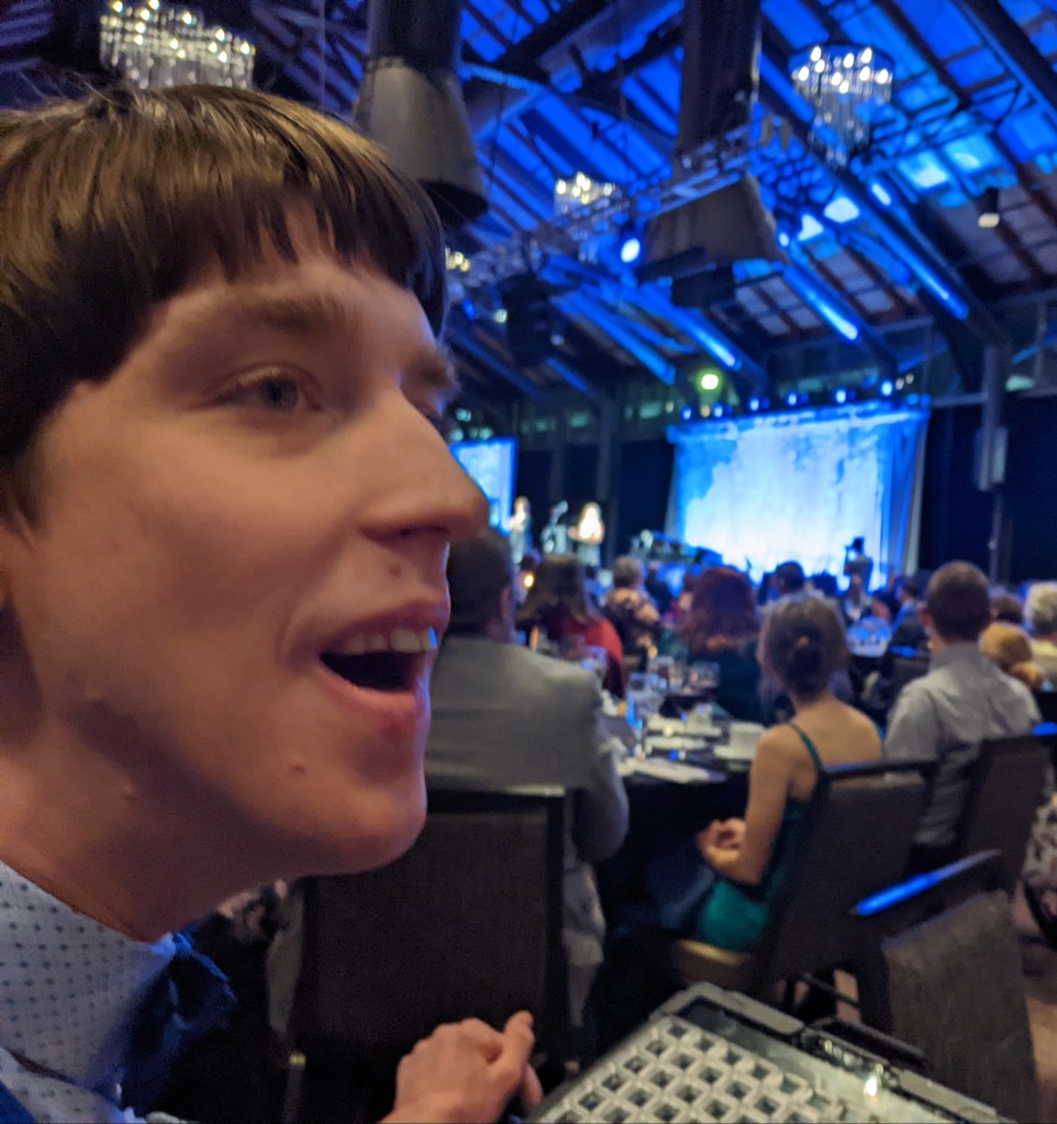 Justin in ballroom with people seated at tables, speaker on stage