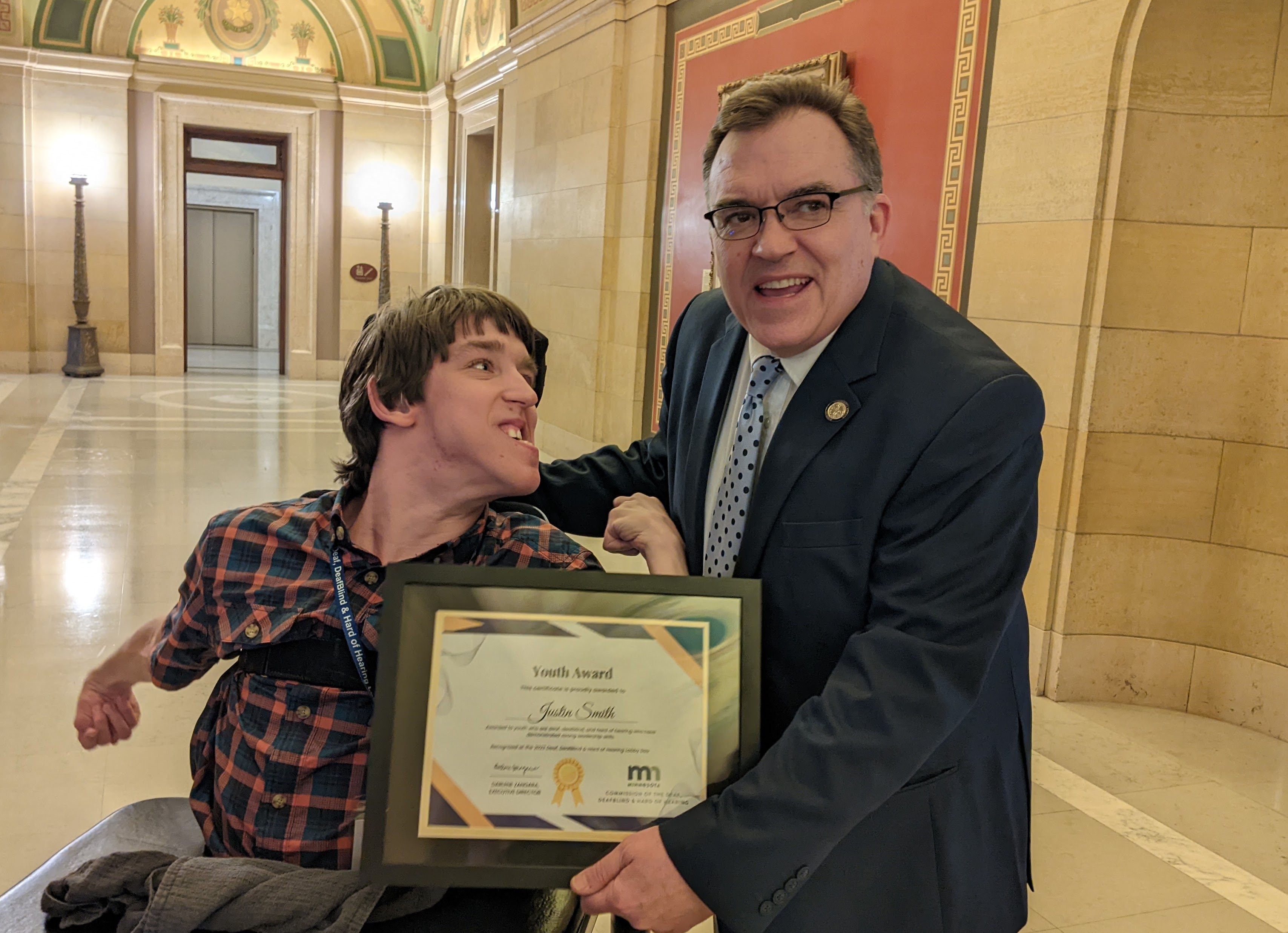Justin seated in wheelchair, looking at Senator John Hoffman who is wearing a suit and holding Justin's Youth Award frame in front of Justin in empty capitol hallway