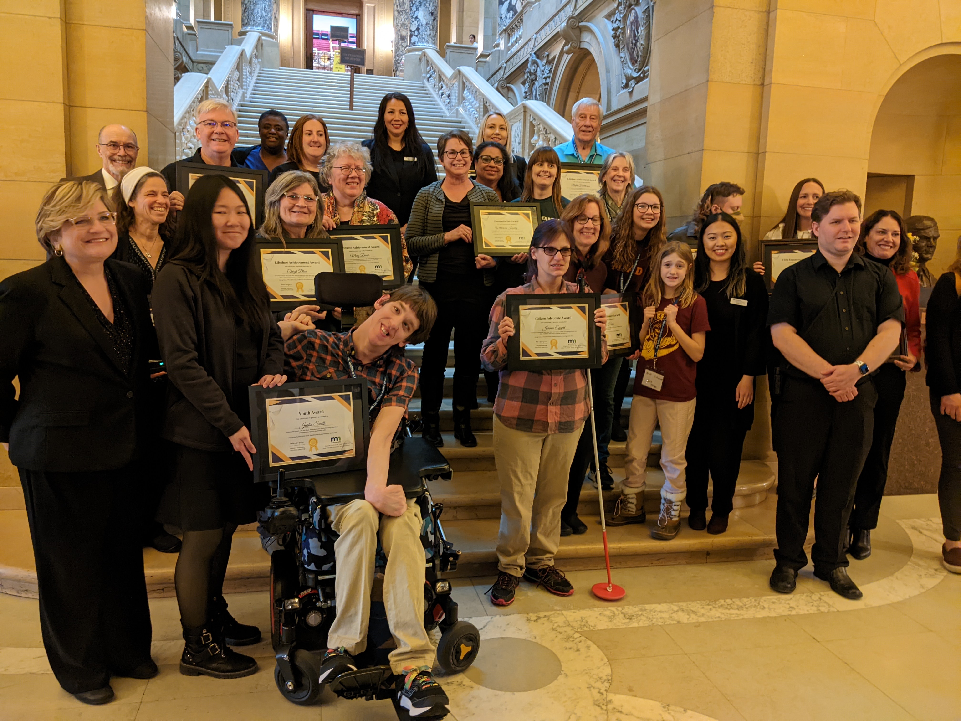 about 25 people standing on stairs inside MN state capitol, smiling and holding award frames