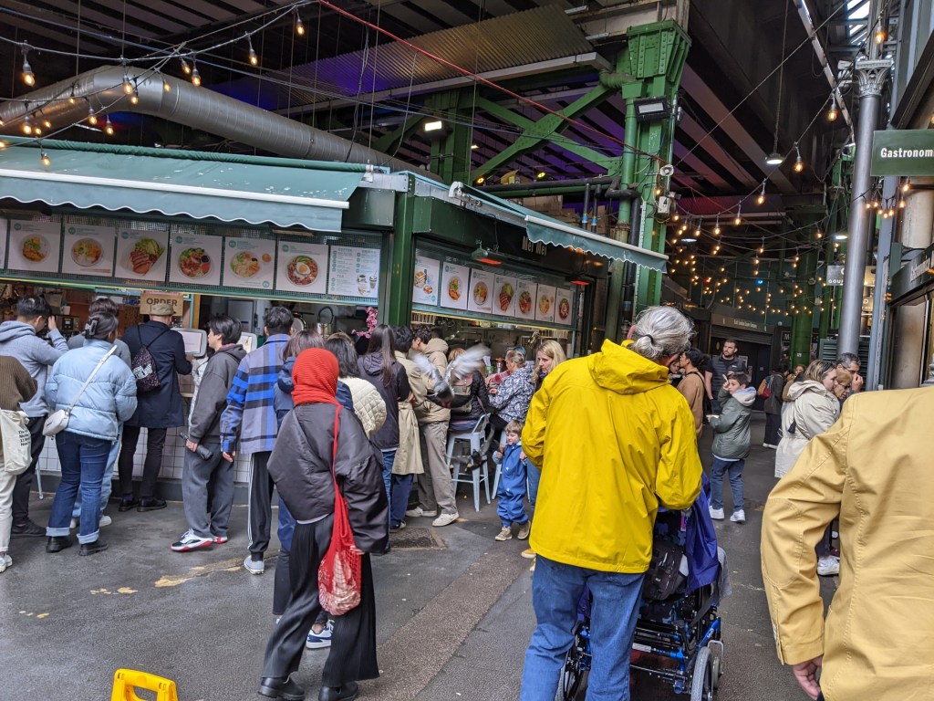 Justin in wheelchair with his dad in yellow rain jacket in borrough market, crowded, food stall
