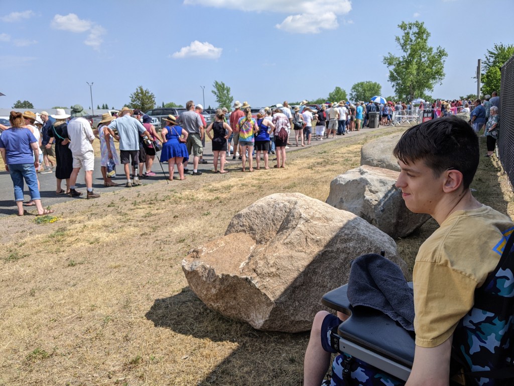 Justin sitting in his wheelchair in a narrow strip of shade from fence behind him, giant boulders, people standing in line outside, brown grass