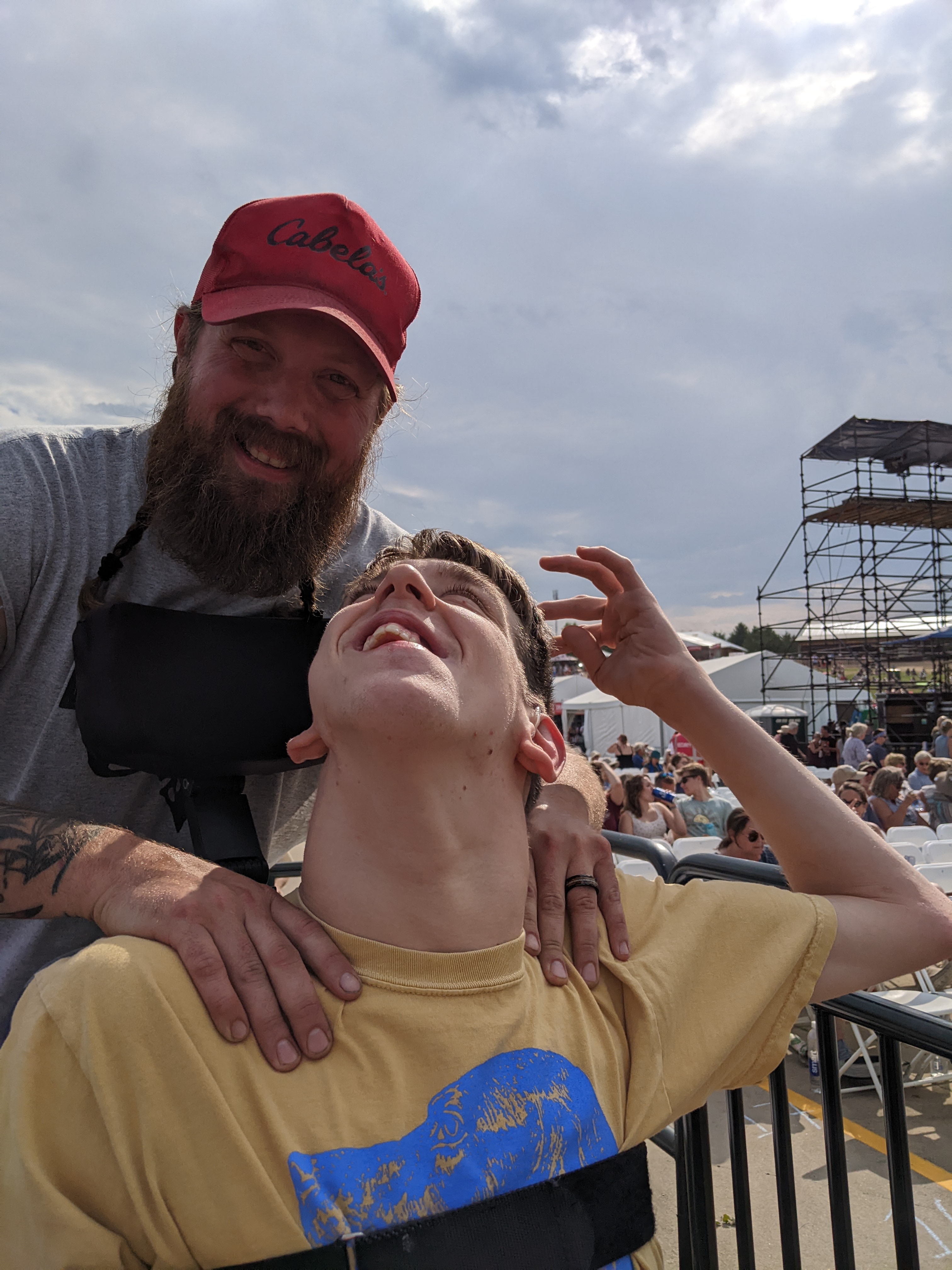 Justin looking up and back towards smiling young man with beard and cap, seated outdoor audience and chairs in background