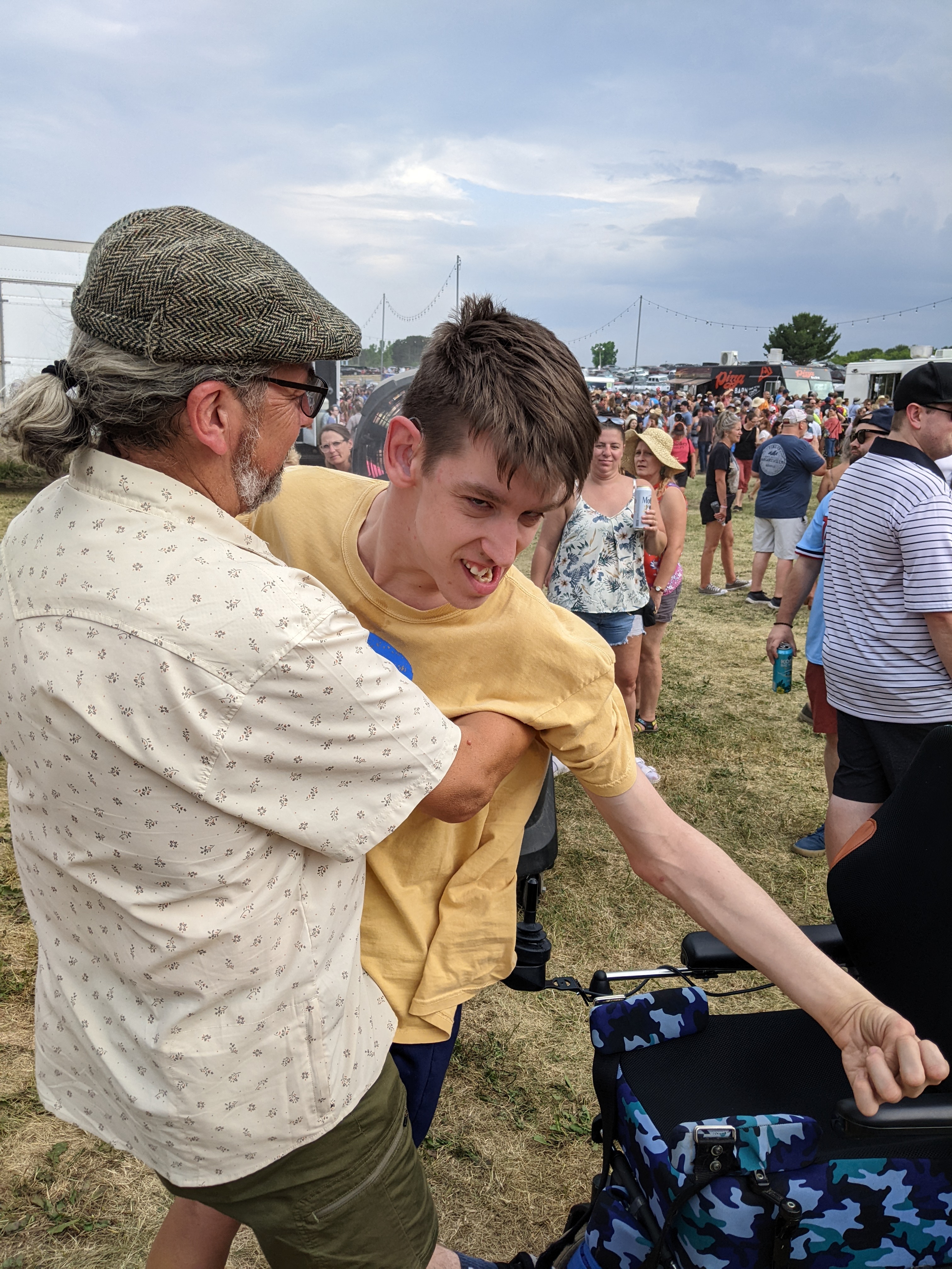 Justin's dad holding him in standing position next to wheelchair, misting fan behind, crowd of people, outdoors