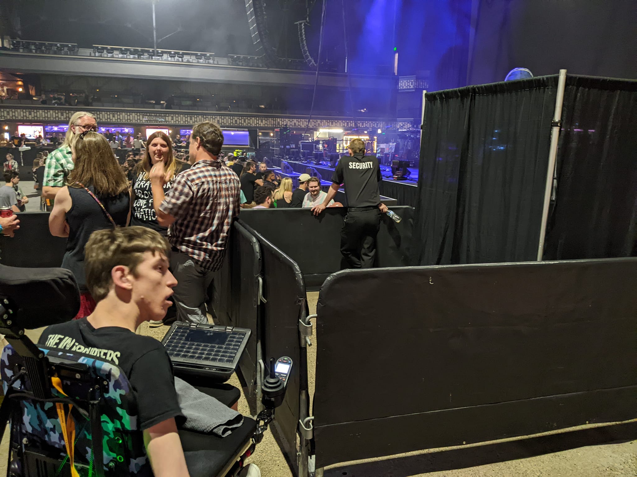 Justin not smiling, sitting in wheelchair, gated off area to the right with man in security shirt, people standing in front of Justin, crowd on main floor in front of stage
