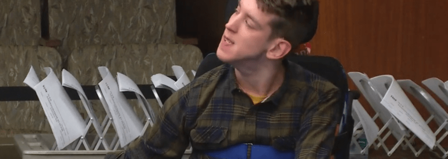 Justin, young man seated in wheelchair, communication device in front of him, looking to the side as he testifies in House hearing room
