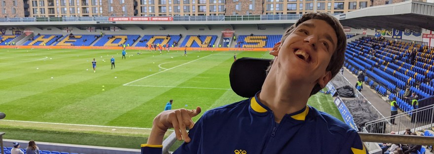 Justin smiling wearing a blue and yellow AFC Wimbledon jacket, Wimbledon football stadium behind with players warming up on the pitch