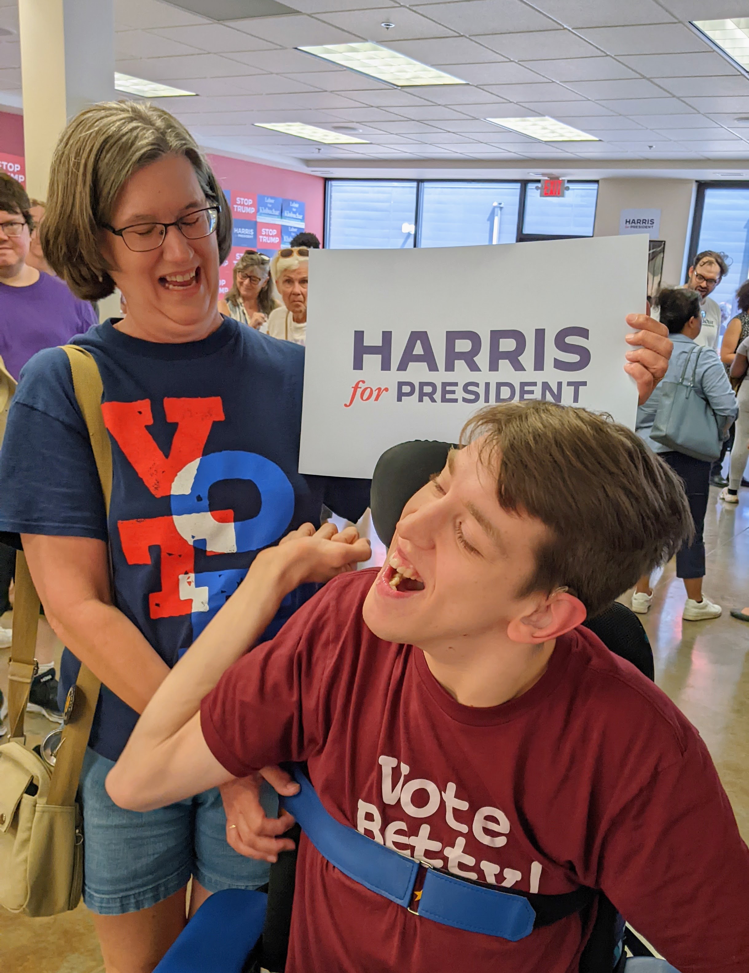 Justin in wheelchair with Vote Betty shirt, his mom next to him with Vote t-shirt holding Harris for President sign