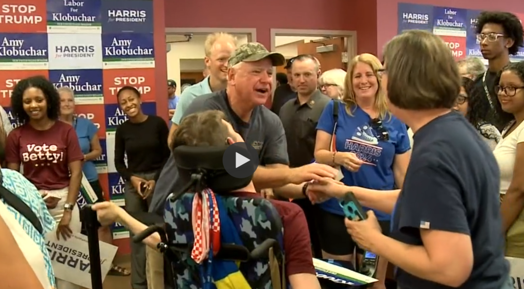 Governor Walz reaching out to shake woman's hand, young man in wheelchair looking at the Governor, crowd smiling on, political posters for Harris, Klobuchar, and stop Trump posted on walls 