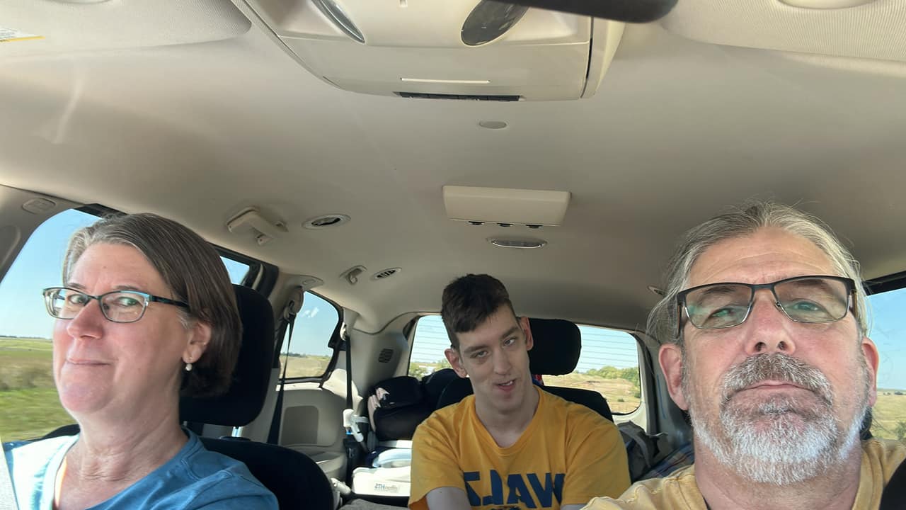 Middle-aged man and woman in front seats of minivan and Justin wearing Walz shirt in wheelchair in middle row, Nebraska prairie out the windows