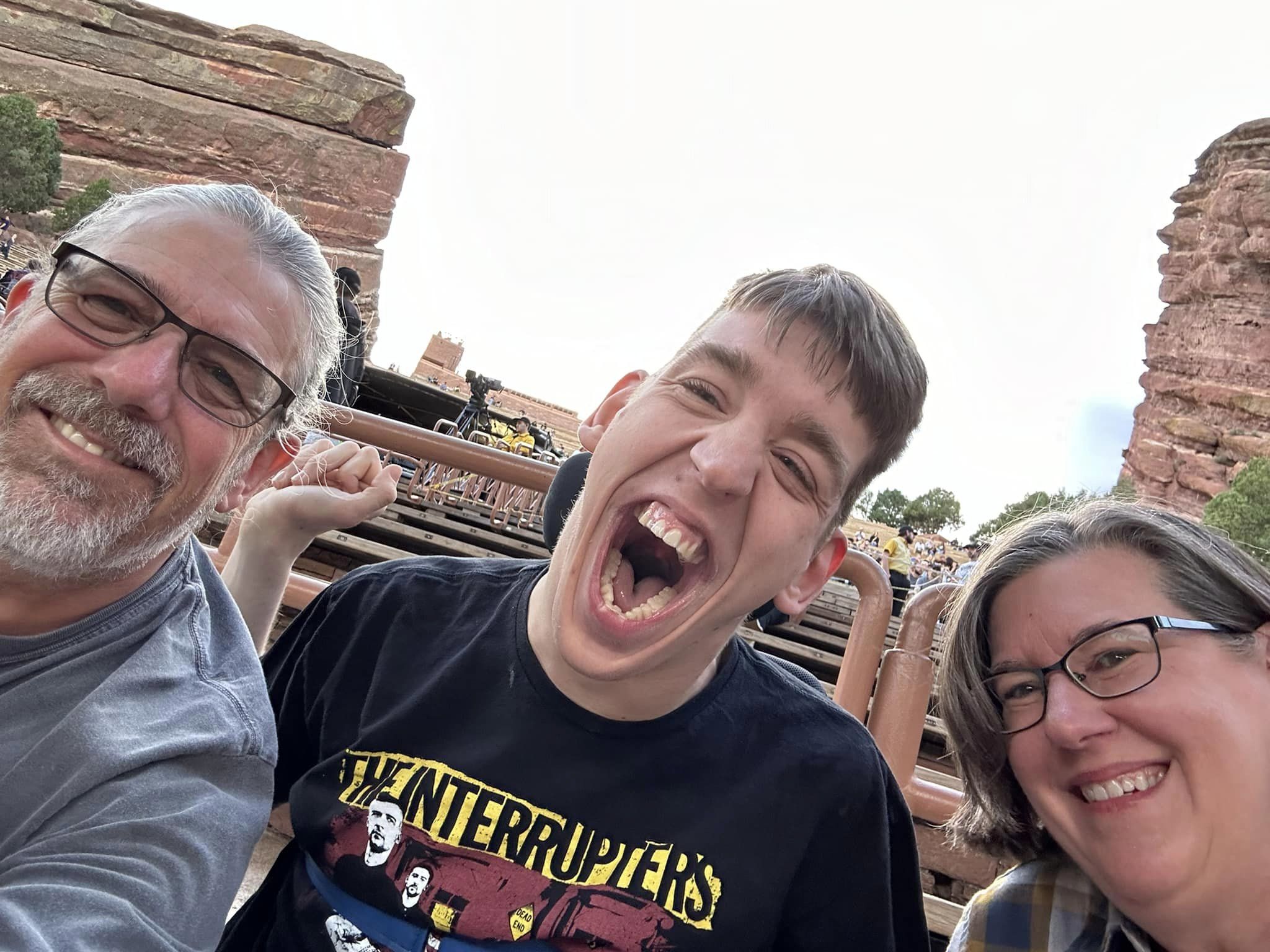 middle aged man and woman with their mid-20s son in the middle wearing The Interrupters t-shirt, red rocks amphitheater with red rocks and rising bleachers behind