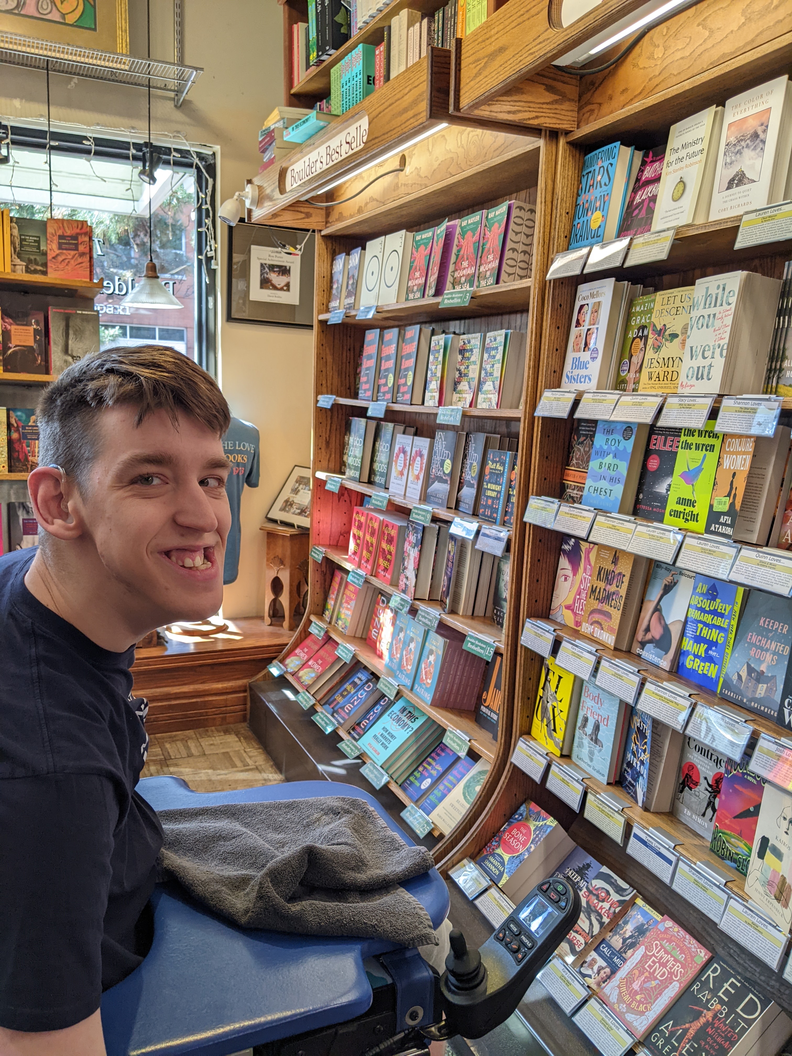 Justin in power wheelchair smiling, in bookstore with lots of books on shelves