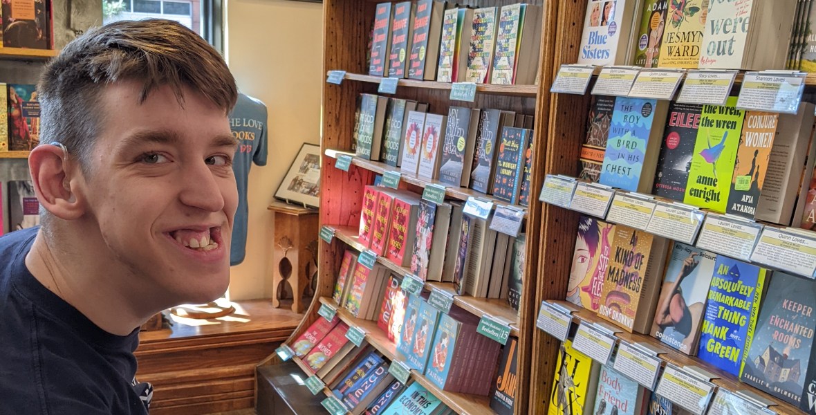 Justin in power wheelchair smiling, in bookstore with lots of books on shelves