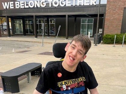 Justin smiling, seated in wheelchair, I voted sticker on shirt,, building with We Belong Together sign behind him